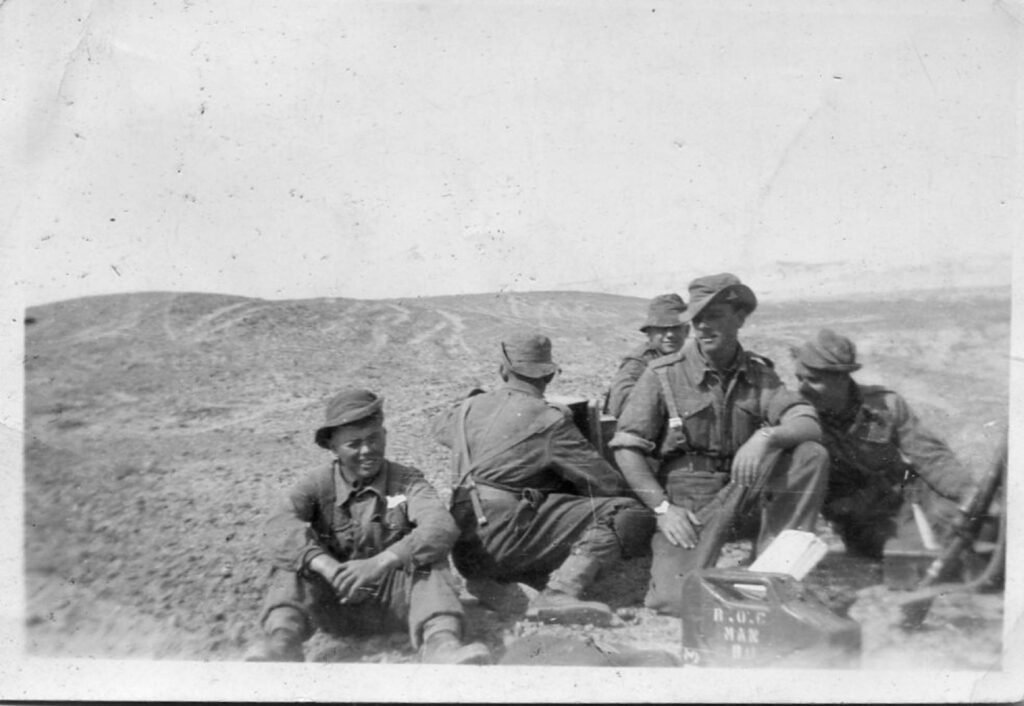 A group of soldiers in a black and white photograph from World War II, outdoors on rugged terrain.