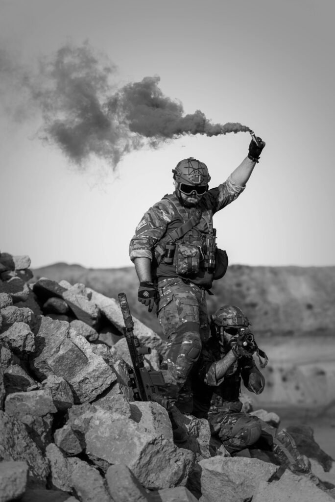 Two soldiers in action with smoke signal in rocky desert terrain.