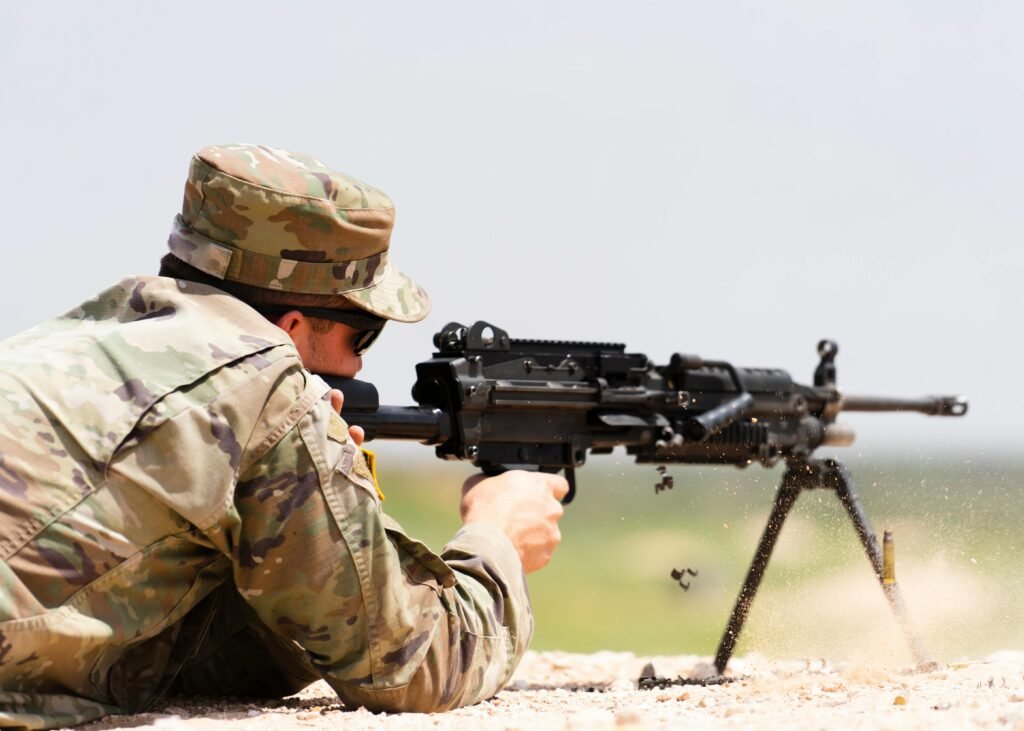 A soldier in camouflage uniform fires an M249 machine gun in an outdoor setting.